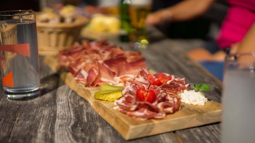Familienhotel in Sterzing: hoi und herzlich willkommen! Brett mit Aufschnitt, Essiggurken, Tomaten und Käse auf Holztisch im Freien