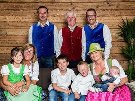 Familienhotel in Sterzing: hoi und herzlich willkommen! Mehrgenerationenfamilie in traditioneller Tracht vor Holzwand