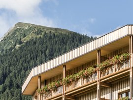 Familienhotel in Sterzing: hoi und herzlich willkommen! Holzhaus mit Balkon und Blumen vor bewaldetem Berg unter blauem Himmel
