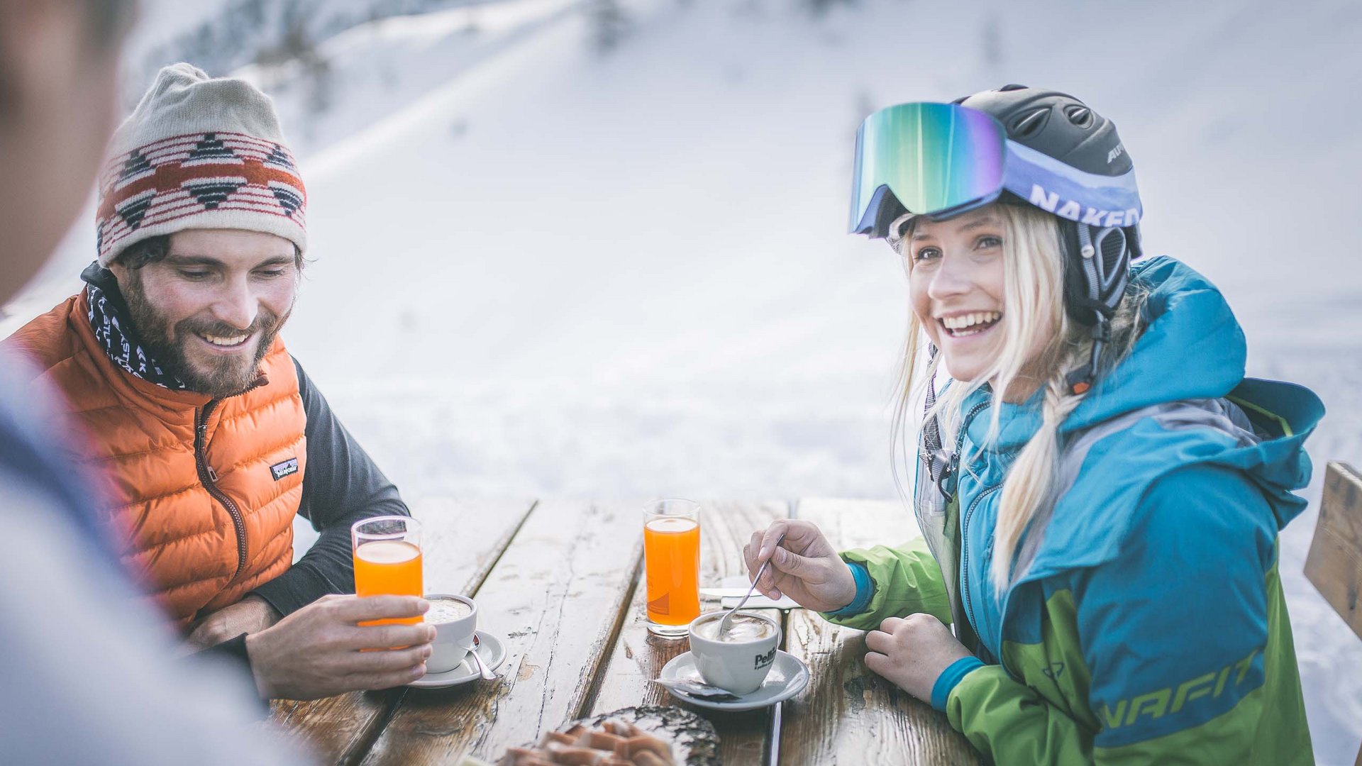 Hotel in Ladurns – perfekt für den Winterurlaub! Zwei Personen trinken Kaffee und Saft an einem verschneiten Außenholzttisch