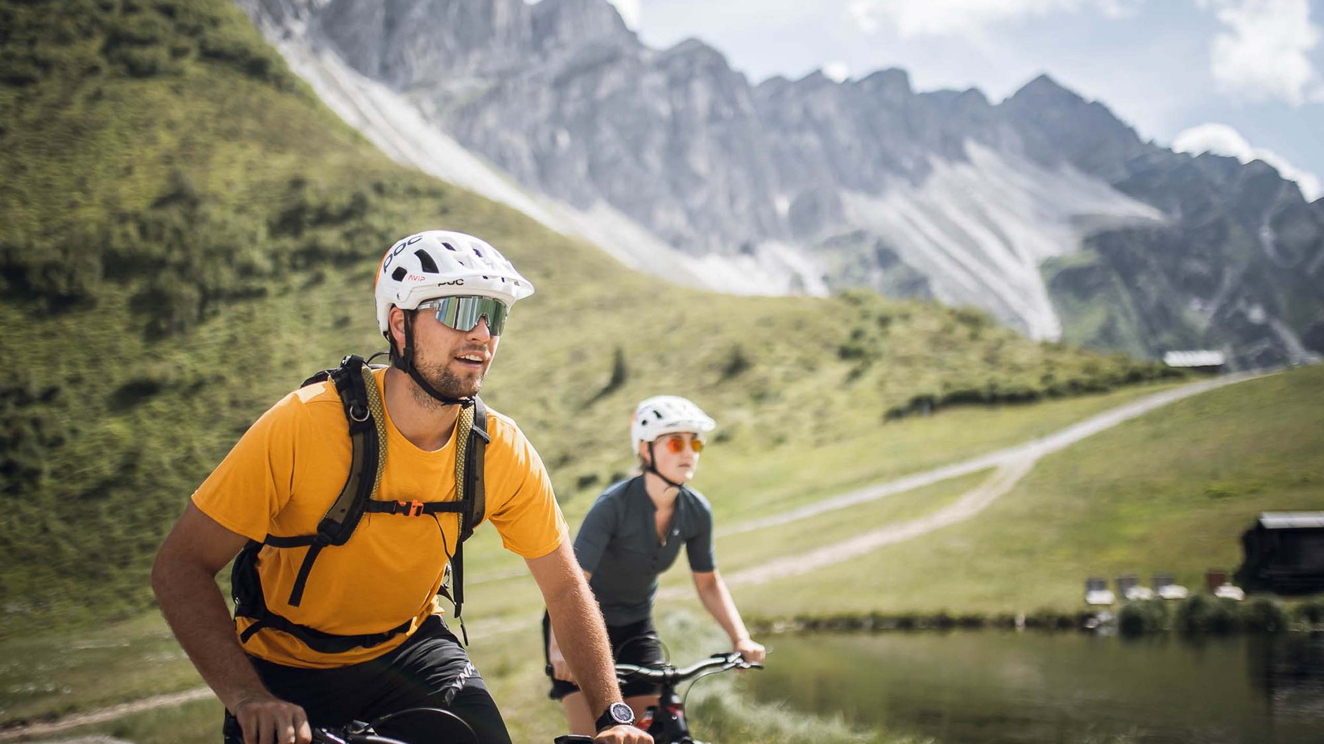 Pflerschtal: accommodation for biking holidays Two cyclists wearing helmets ride on a mountain path beside a lake