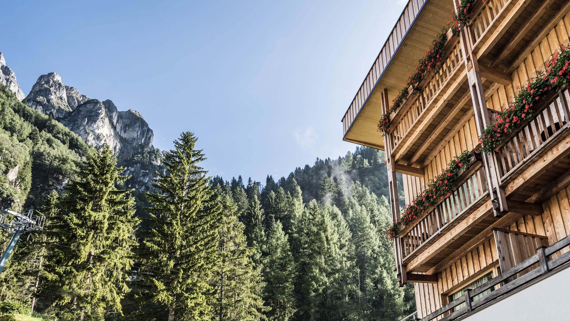 Familienhotel in Sterzing: hoi und herzlich willkommen! Holzhaus mit Balkonen und Blumen neben Tannen und Bergen unter blauem Himmel