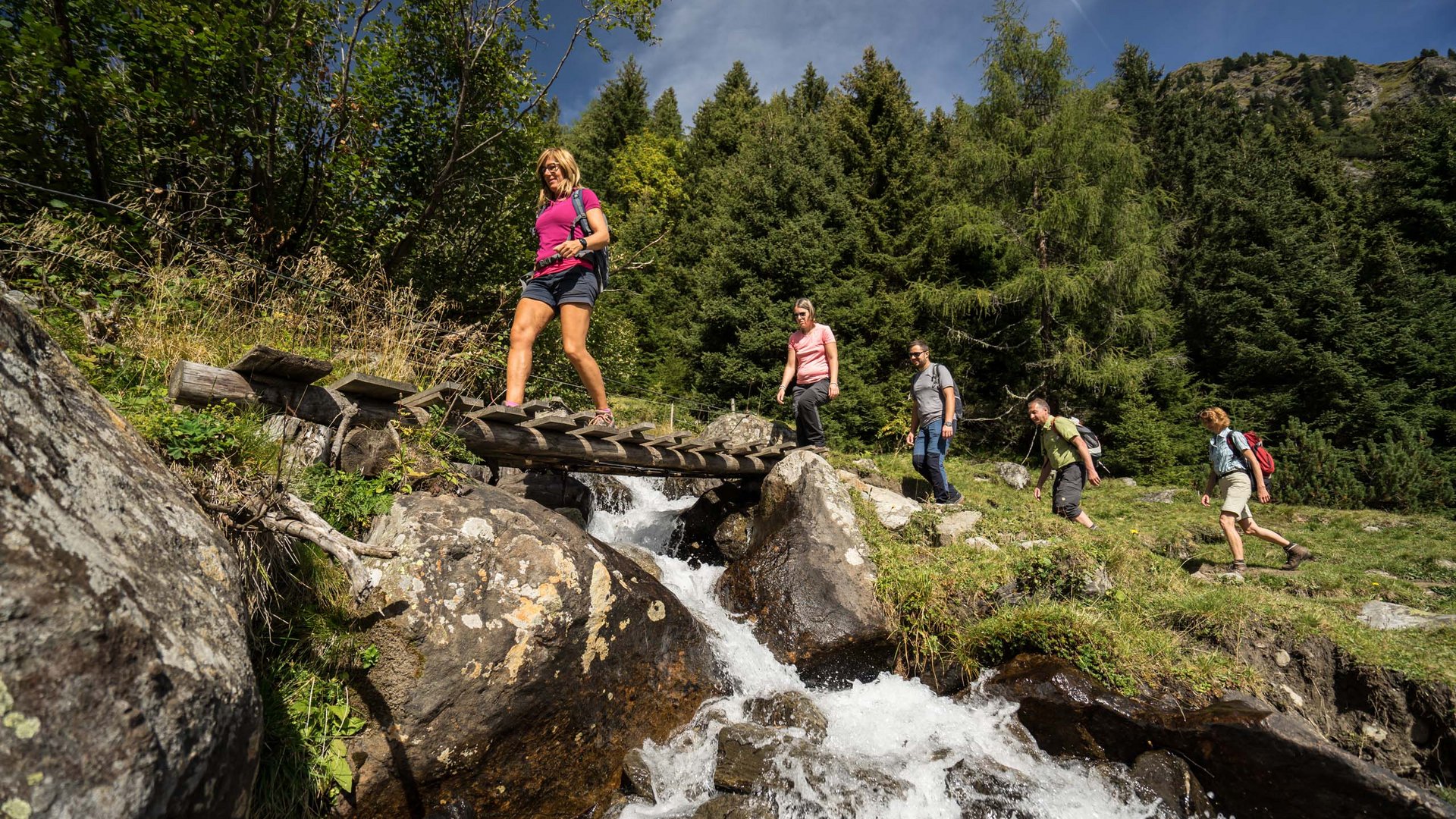Familienhotel in Sterzing: hoi und herzlich willkommen! Gruppe wandert über kleine Holzbrücke an Bergbach im Wald